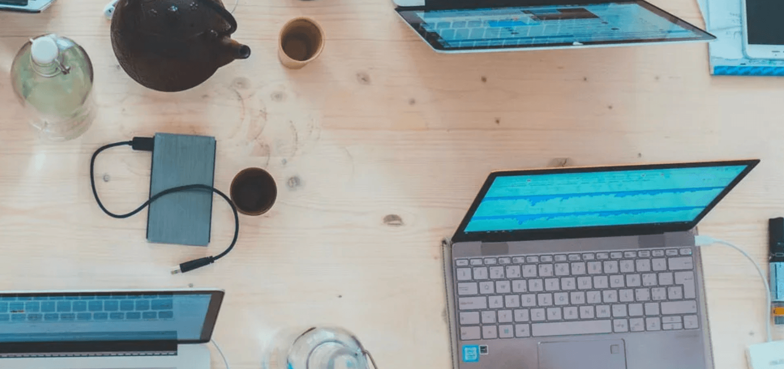 Flat lay of multiple laptops, a power bank, and tea cups on a light wooden desk during a collaborative workspace session.